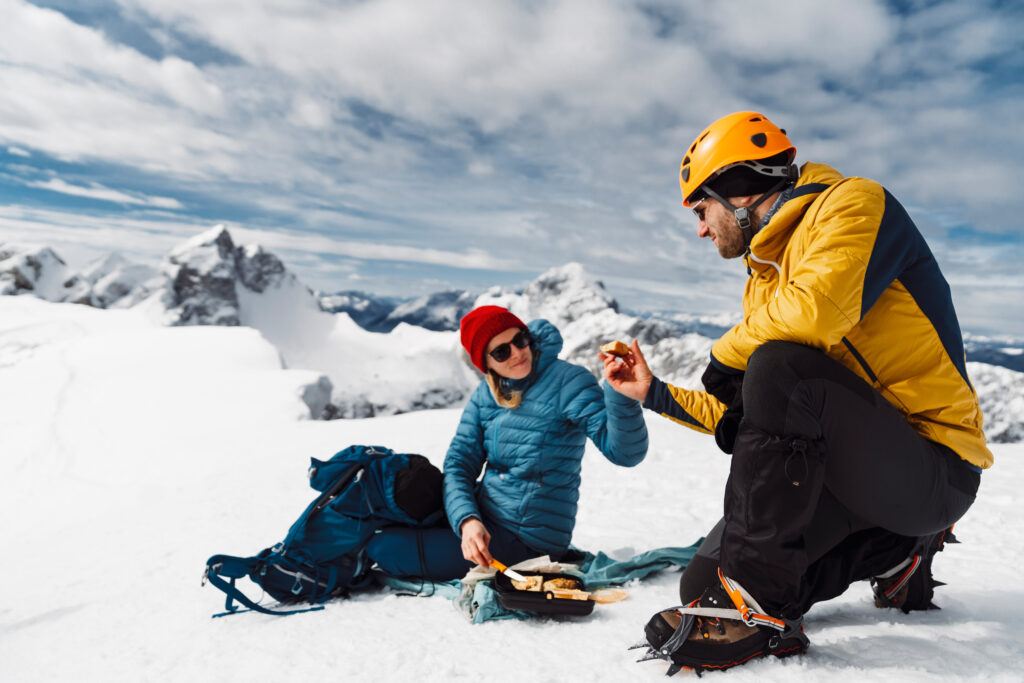 Mountaineering couple having lunch on top of the snowy mountain, sitting on the sun in the snow